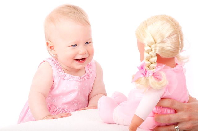 A photograph of a baby in a pink dress smiling at a doll.