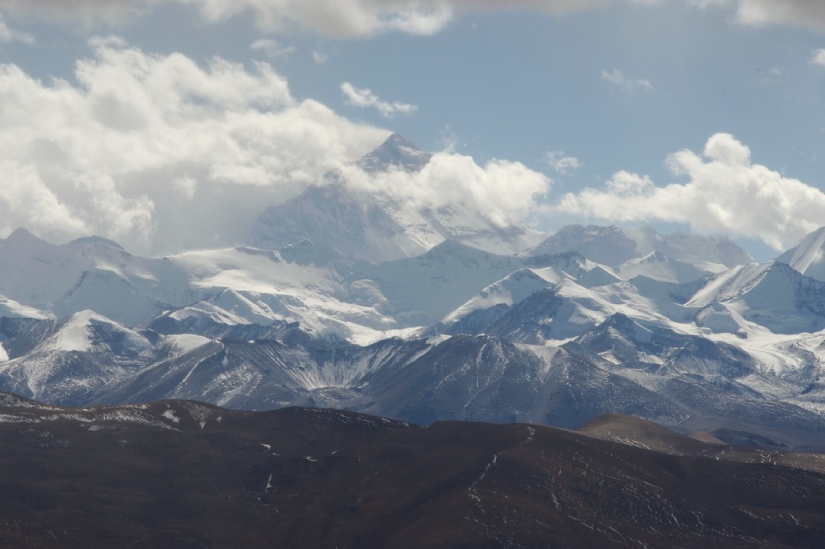 This photo shows Mount Everest as seen from a distance. It is a large, pyramid-shaped, craggy peak with many smaller snow-covered peaks in the foreground. The peak of Mount Everest is partially occluded by clouds.