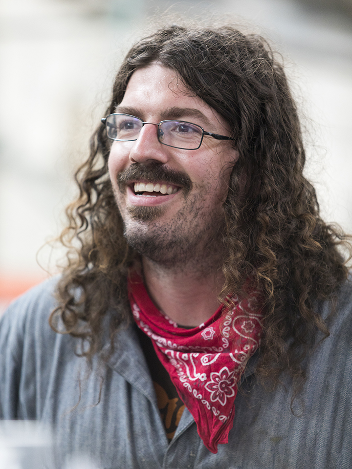 Photo of a smiling bearded man with long curly brown hair wearing blue coveralls and a red scarf around his neck.